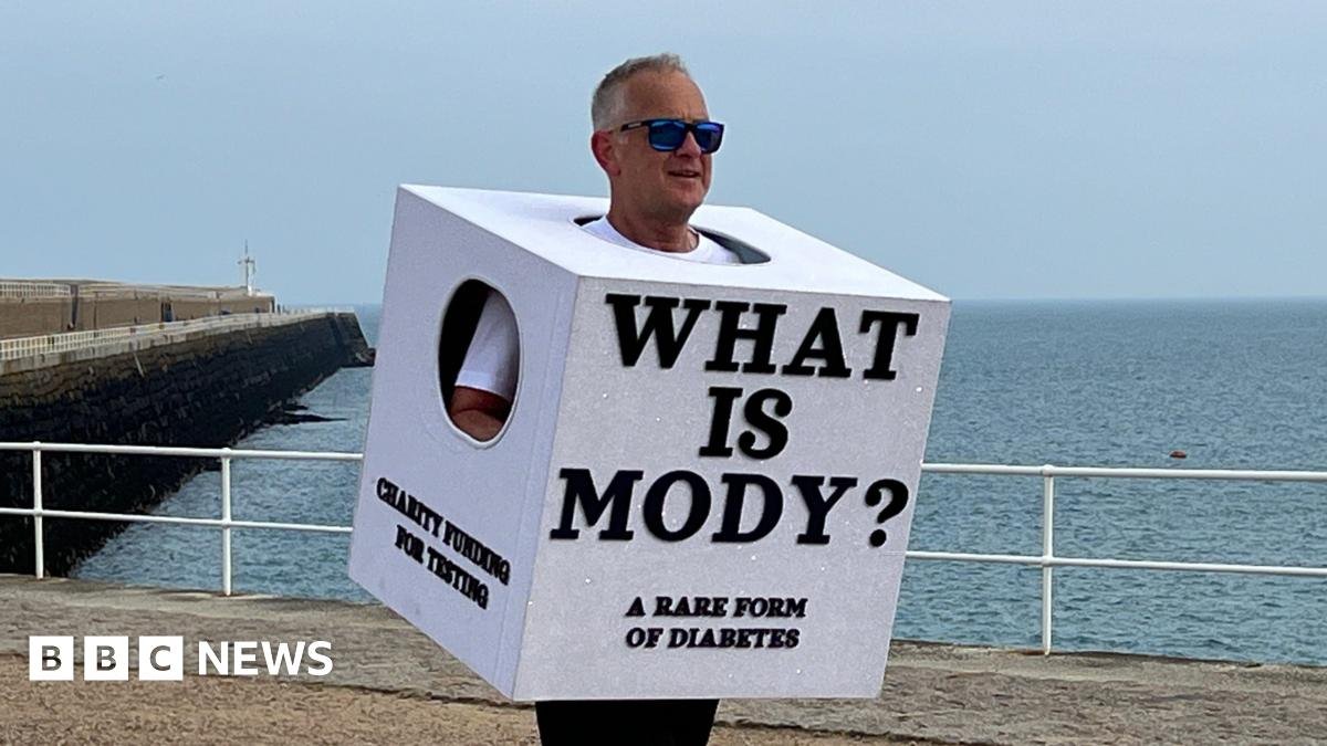 Richard is running long a seaside promenade wearing a large white cube costume with the text ‘WHAT IS MODY? A RARE FORM OF DIABETES,’ with the ocean and a pier behind him
